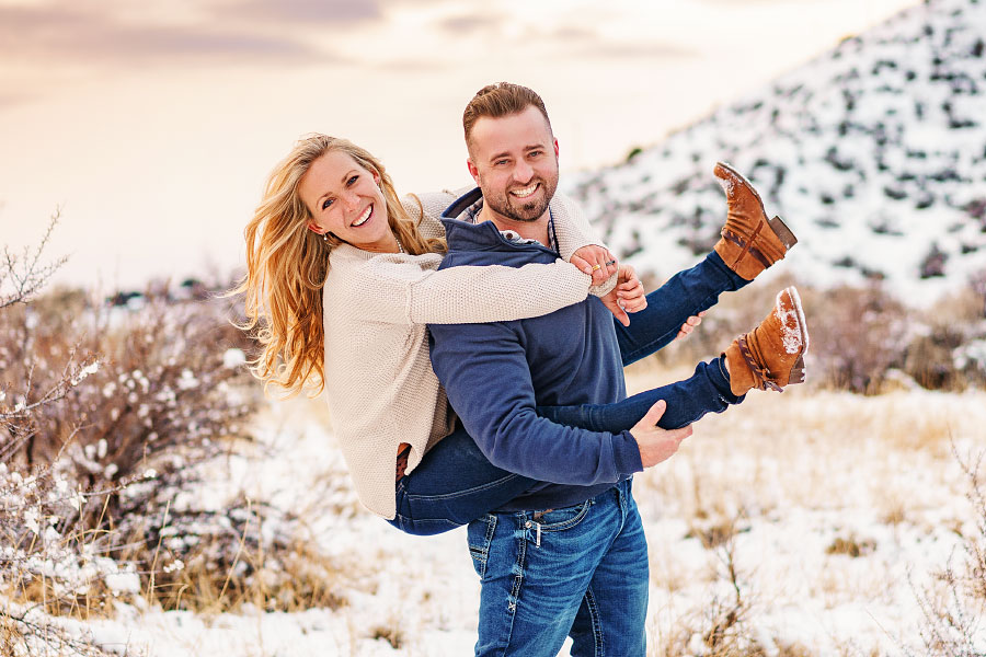 Laughing man gives woman a piggyback ride for couples session in snowy Boise Idaho foothills