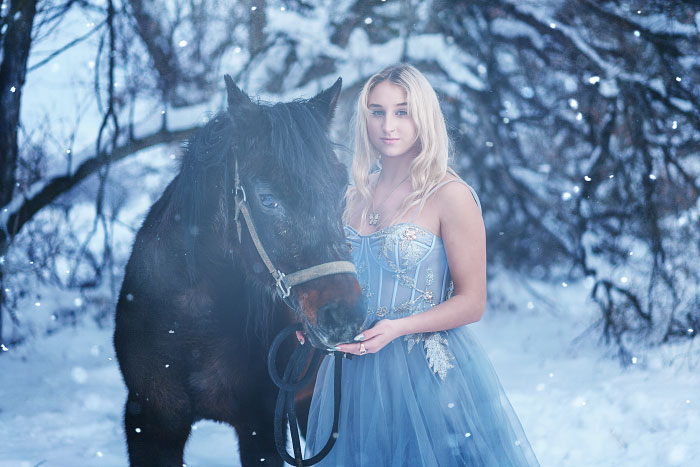 High school senior girl in blue dress stands by horse in snowy landscape for senior photos in Idaho