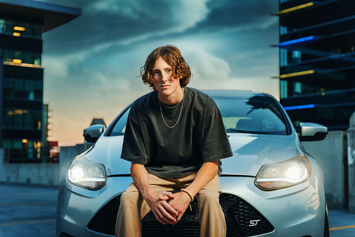 Young man sits on hood of car atop parking garage in downtown Boise for senior photo session
