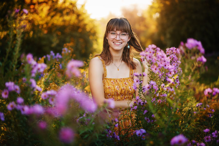Brunette teen girl with glasses wearing yellow sun dress stands in meadow of purple flowers for senior photoshoot
