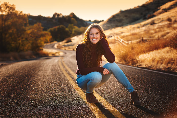 Senior girl squats on two-lane country road for senior photos