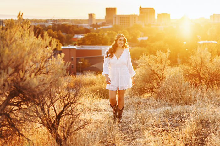 Teen girl wearing white dress walks through golden field in Boise foothills at sunset for her senior photo session