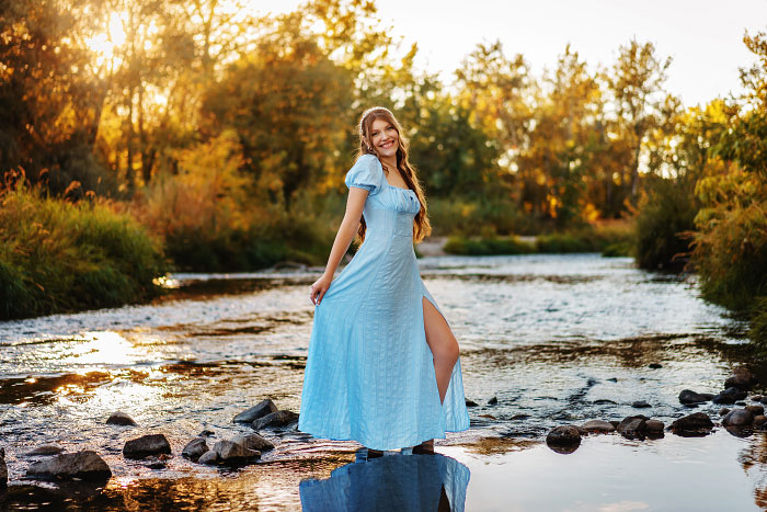 Teen girl wearing flowing blue dress stands in Boise River for senior photos in Eagle, Idaho