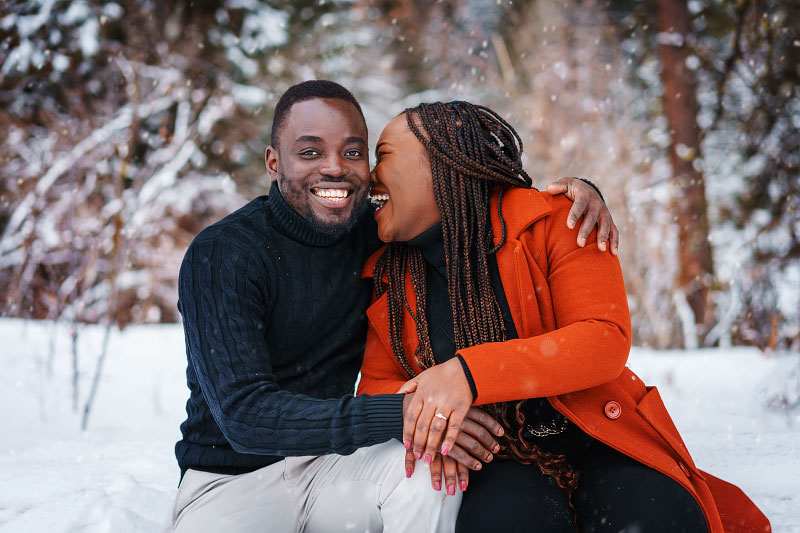 Young Black couple laughs together in snow, showing off new engagement ring for portrait session