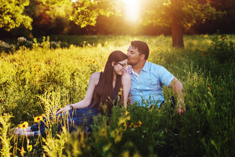 Young man kisses young woman's forehead sitting in sunlit meadow in Boise's Kathryn Albertson Park for couples session