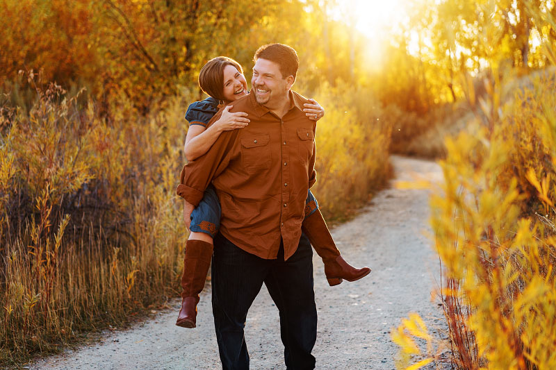 Man gives woman a piggyback ride down gravel hiking trail at Military Reserve Park in Idaho in autumn