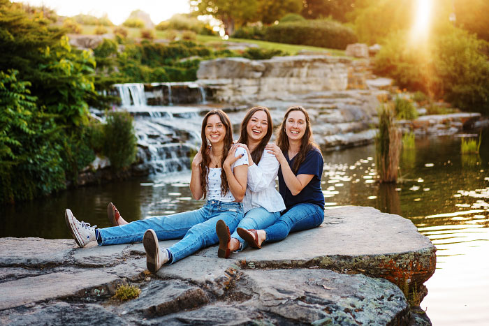 Three adult sisters sit in line laughing at Boise depot grounds with waterfalls in background