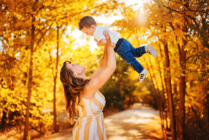 Laughing mom holds young son up high Superman-style on tree-lined autumn path in Eagle Idaho