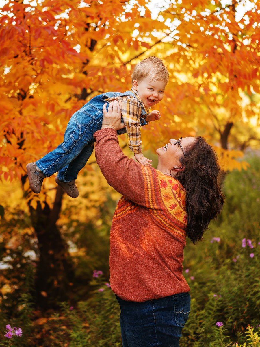 Mom holds laughing young son over head with background of vibrant orange trees at Boise's Kathryn Albertson Park